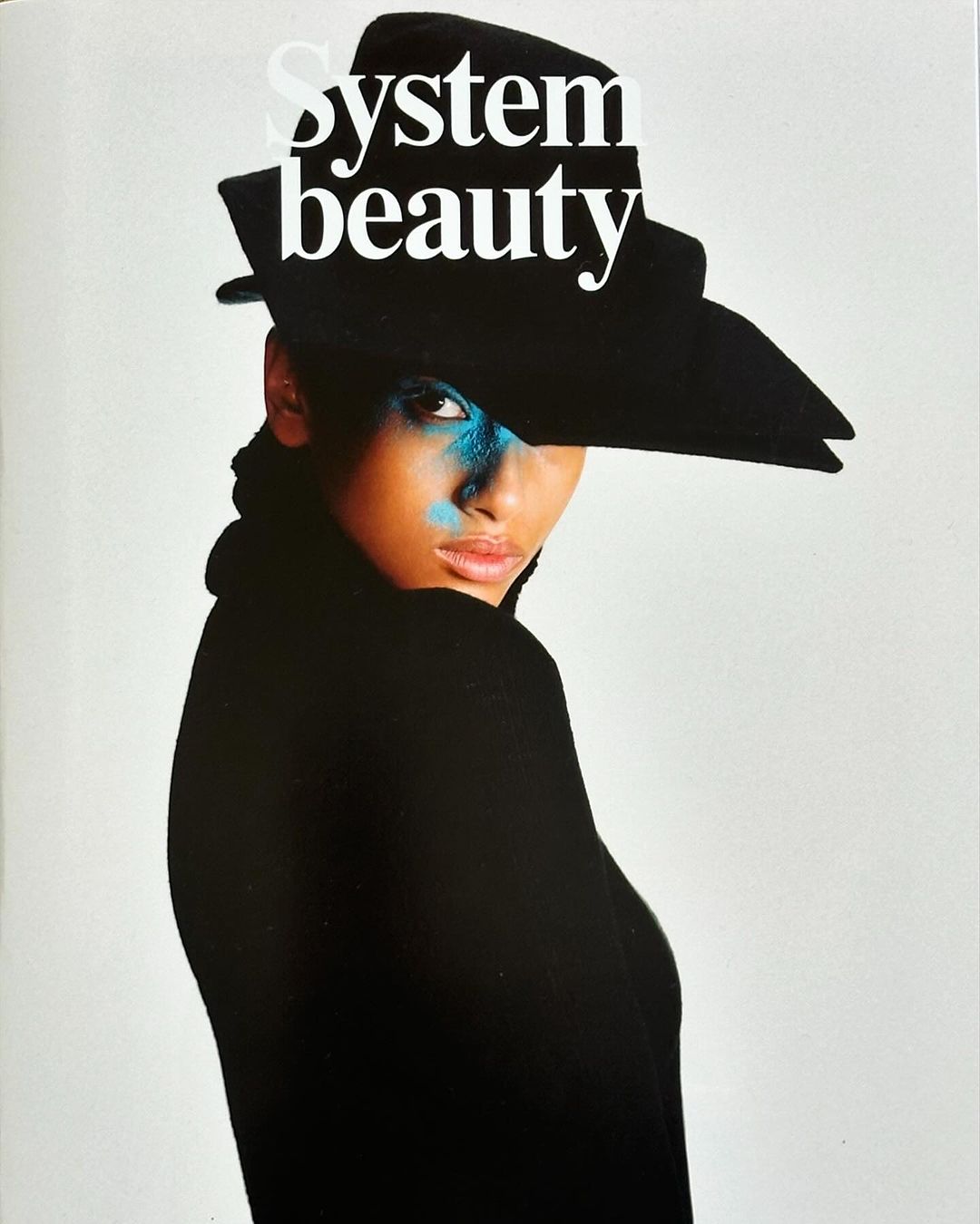 Woman wearing black hat and blue eye makeup, posing against white backdrop, editorial style fashion portrait for magazine cover.