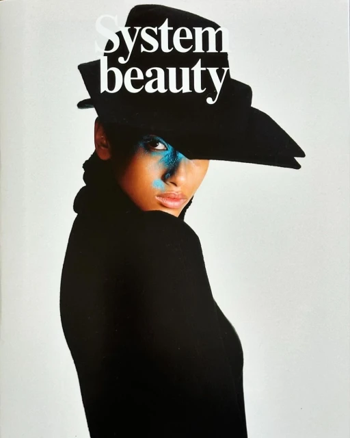 Woman wearing black hat and blue eye makeup, posing against white backdrop, editorial style fashion portrait for magazine cover.