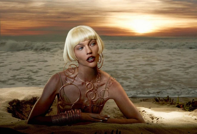 Model with blonde bob wig and large gold hoop earrings posing on a sandy beach at sunset by Carlijn Jacobs