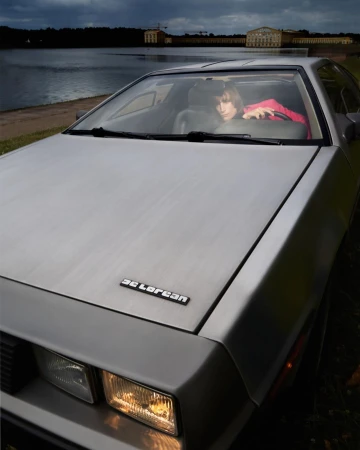 Silver DeLorean car parked near a lake with a building in the distance and a person visible inside the vehicle.