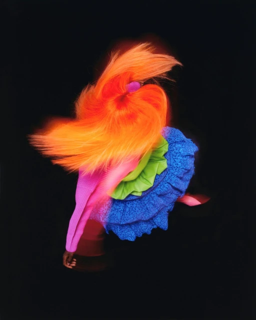 High fashion portrait of a model in motion wearing a ruffled dress with neon orange hair against a dark studio backdrop.