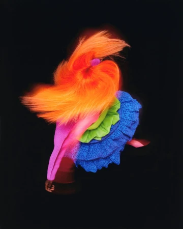 High fashion portrait of a model in motion wearing a ruffled dress with neon orange hair against a dark studio backdrop.