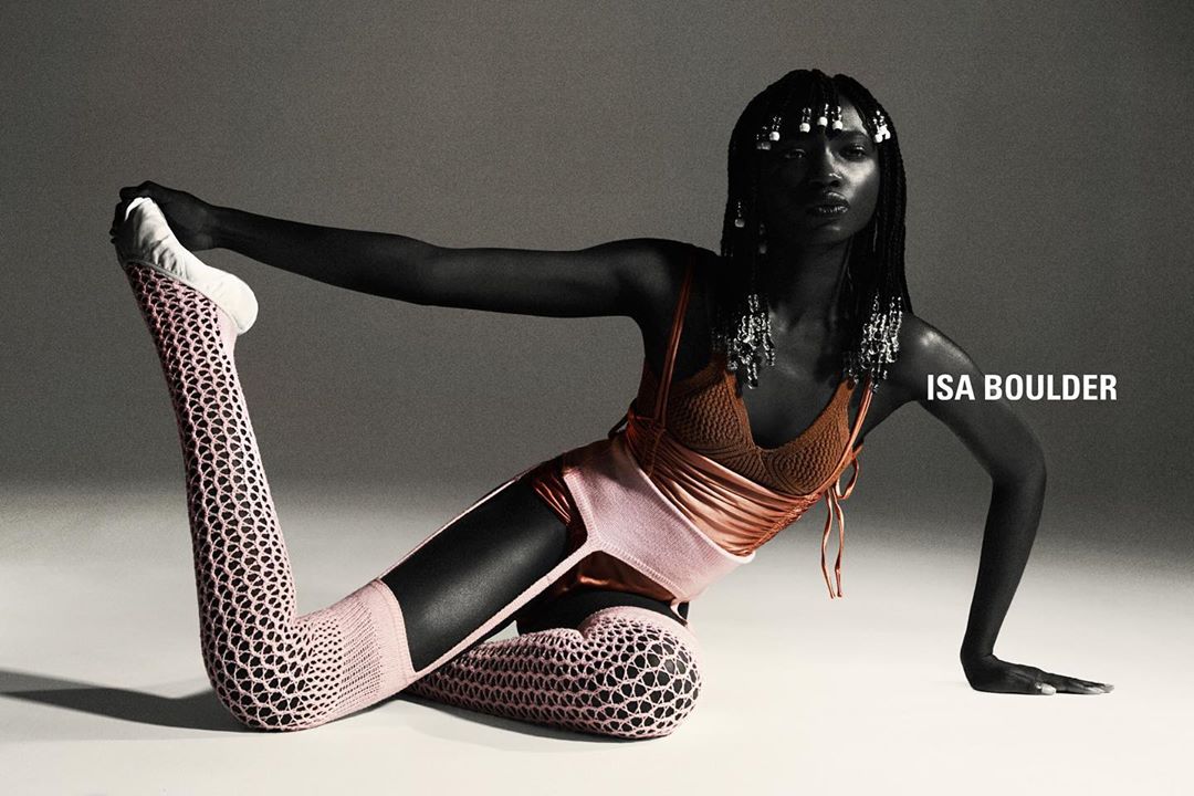 An African-American woman in artistic yoga pose wears a brown top, peach bottoms and leggings with a cut-out design in a studio shot.