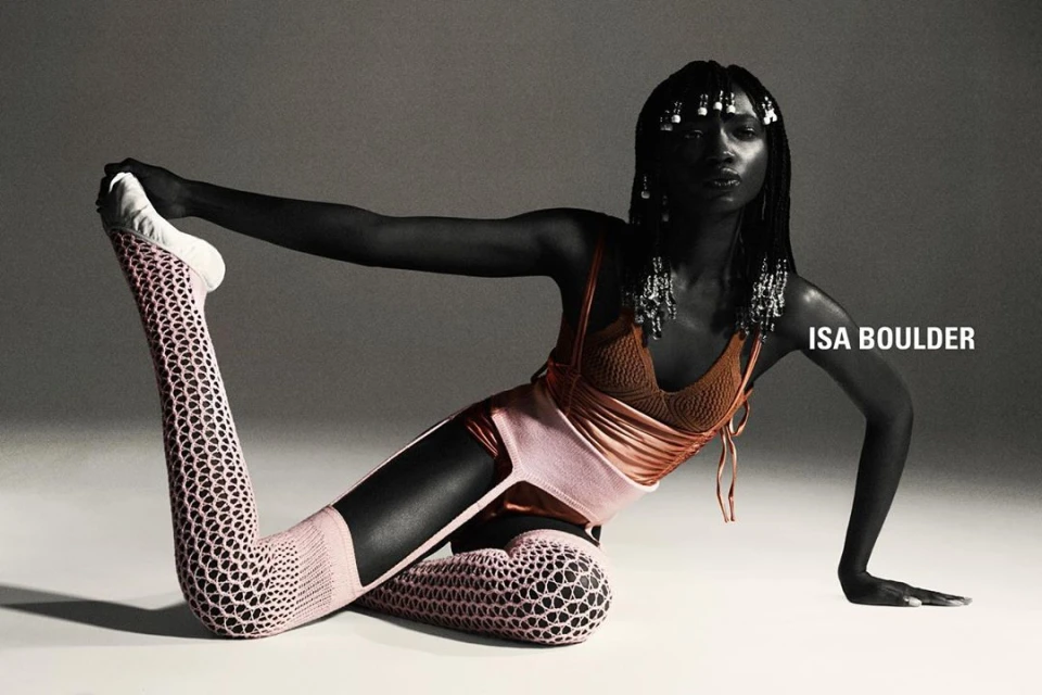 An African-American woman in artistic yoga pose wears a brown top, peach bottoms and leggings with a cut-out design in a studio shot.
