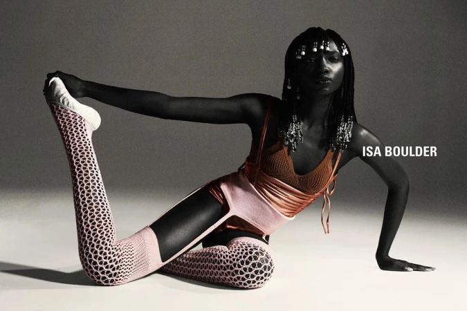 An African-American woman in artistic yoga pose wears a brown top, peach bottoms and leggings with a cut-out design in a studio shot.