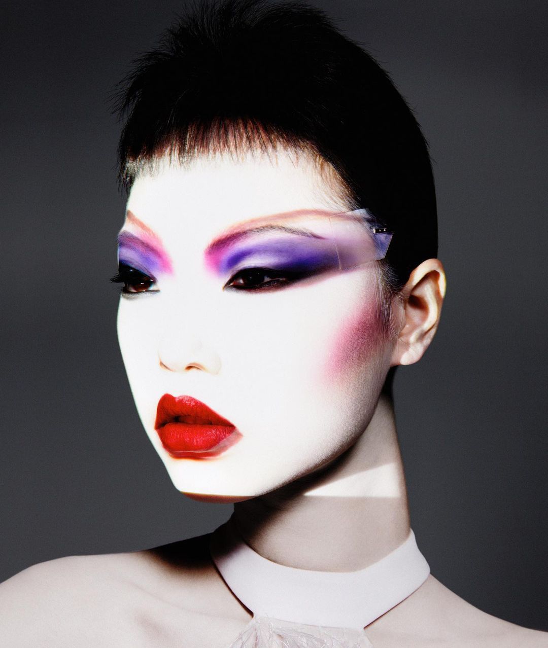 Close-up portrait of a woman with theatrical makeup: bright red lips, purple eyeshadow, white face powder, and short black hair against a gray background.