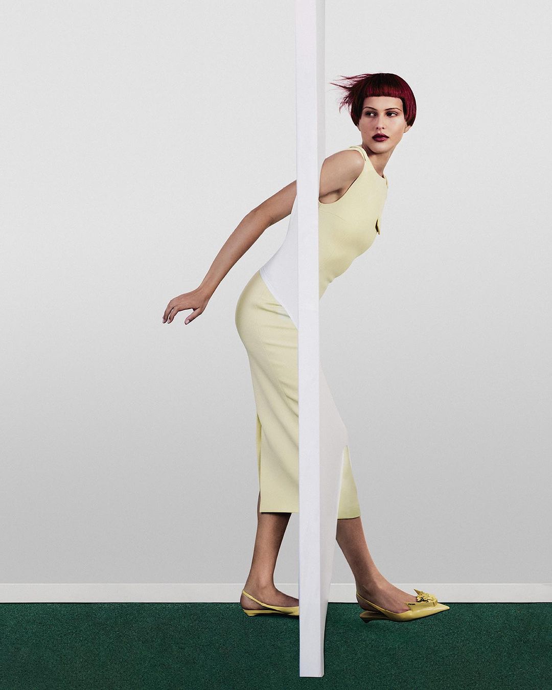 A fair-skinned woman with a red bob, wearing a yellow dress, poses dynamically in front of a white wall with green carpet.