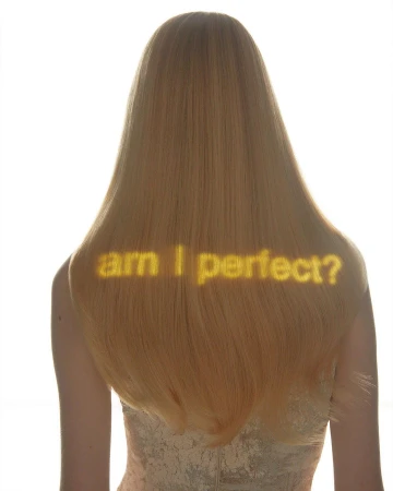 Back view of a fair-skinned adult woman with long blonde hair and the text 'am I perfect?' projected on her back in a bright studio.