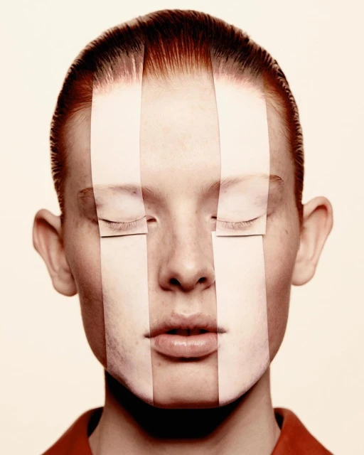 Close-up studio shot of a young adult woman with red hair and pale skin with paper strips obscuring parts of her face.