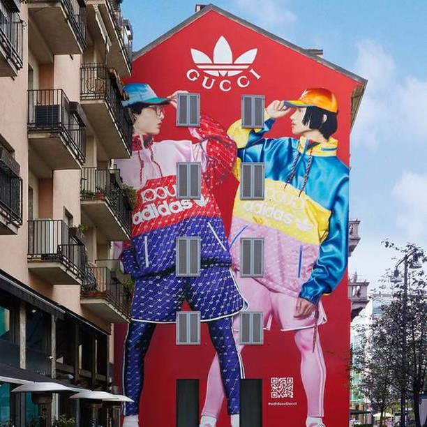 Building mural featuring two people wearing Adidas x Gucci outfits, standing against a red wall with the Gucci logo on an urban street.