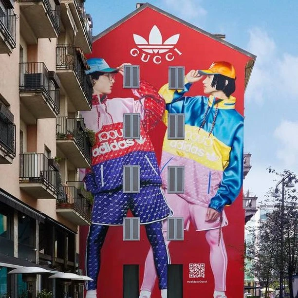 Building mural featuring two people wearing Adidas x Gucci outfits, standing against a red wall with the Gucci logo on an urban street.