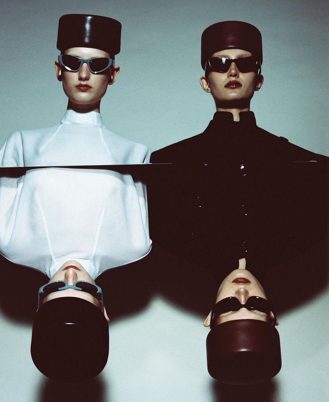Two models wearing structured hats and sunglasses, one in white and one in black, in a mirrored vertical composition by Carlijn Jacobs