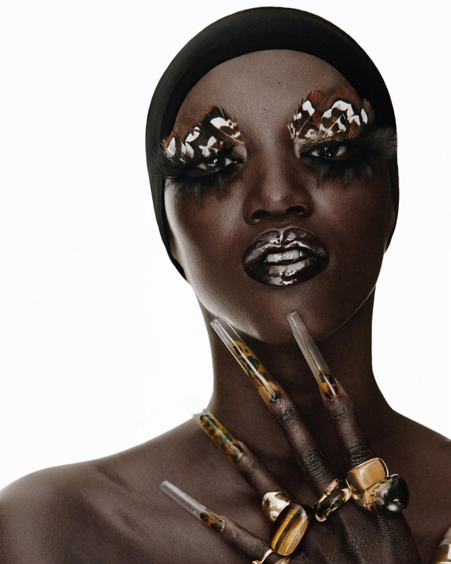 Close-up of a dark-skinned woman with butterfly and feather eye makeup, glossy lips, a black headscarf, and large rings on hands with long nails.