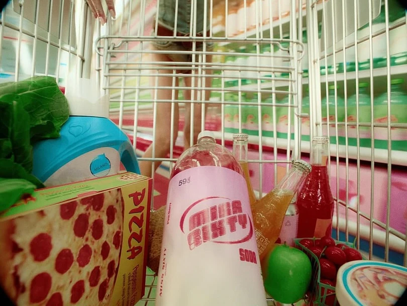 Low-angle view inside a shopping cart filled with groceries: pizza box, soda bottle, fruits, and various beverage bottles.