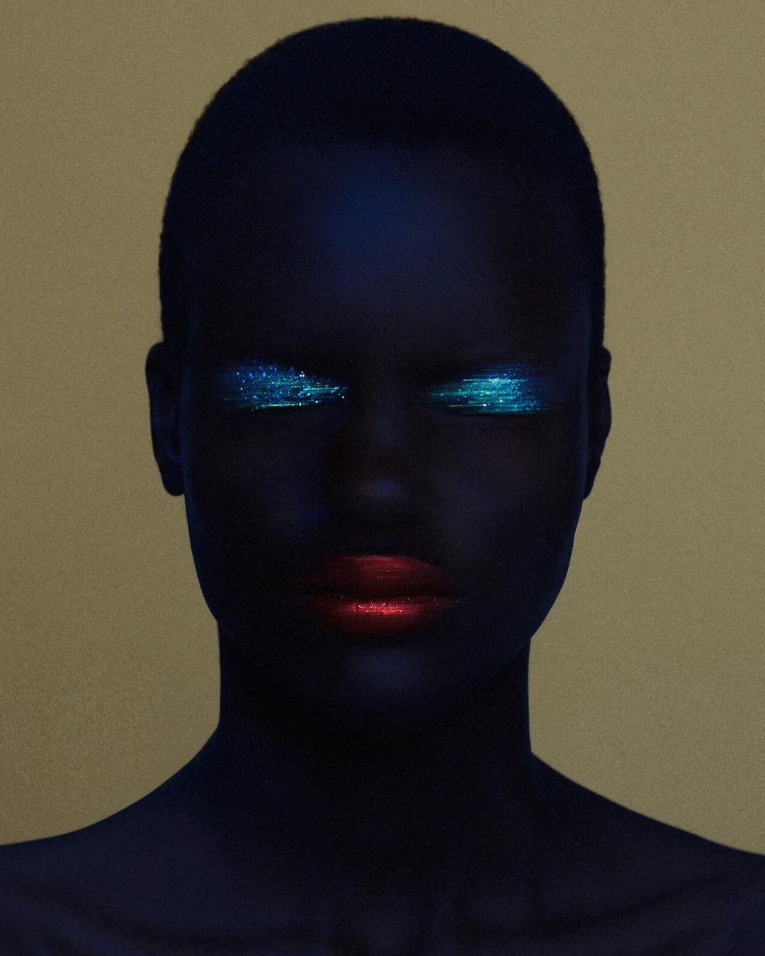 Close-up portrait of a Black woman with dark blue painted skin, glowing turquoise glitter eye makeup, and bold red lips against a gold backdrop.