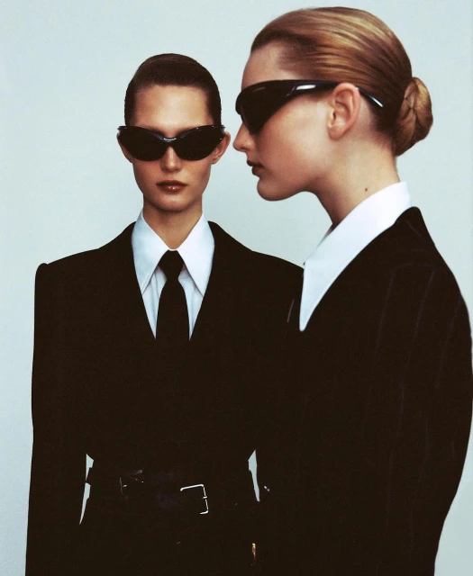Two women with slicked-back hair pose in matching black suits and sunglasses against a plain background in a studio portrait.