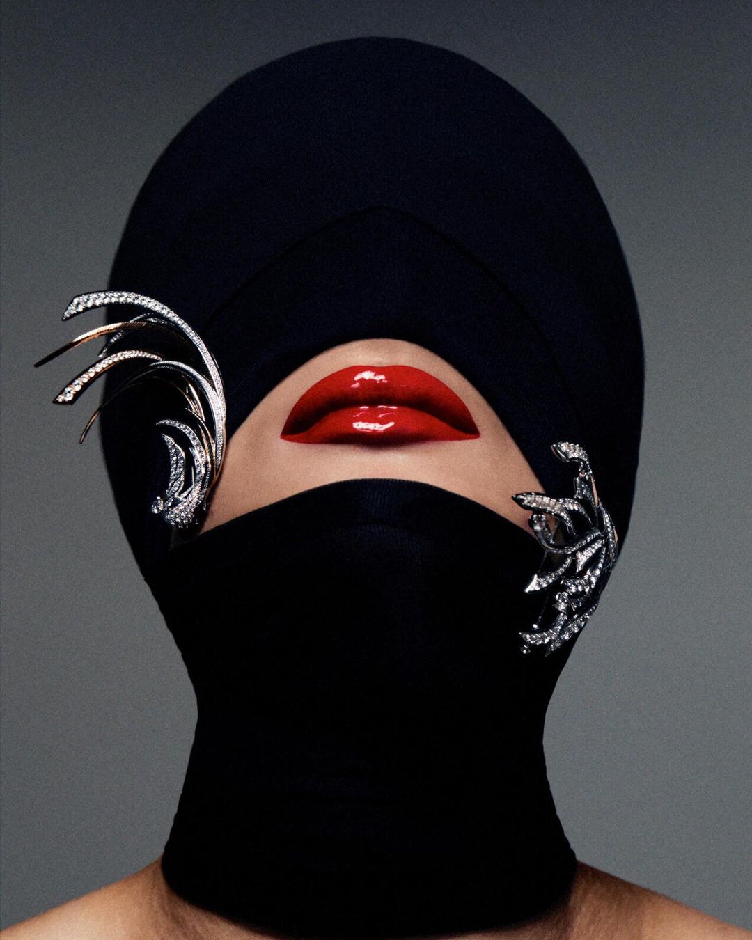High-fashion portrait of a model with bright red lips, wearing a black head covering and jeweled ornamentation against a grey backdrop.