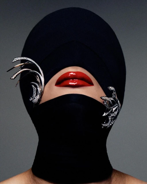 High-fashion portrait of a model with bright red lips, wearing a black head covering and jeweled ornamentation against a grey backdrop.