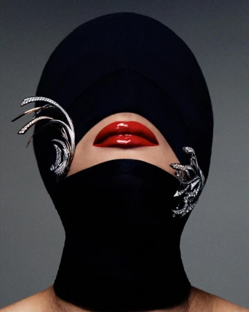 High-fashion portrait of a model with bright red lips, wearing a black head covering and jeweled ornamentation against a grey backdrop.