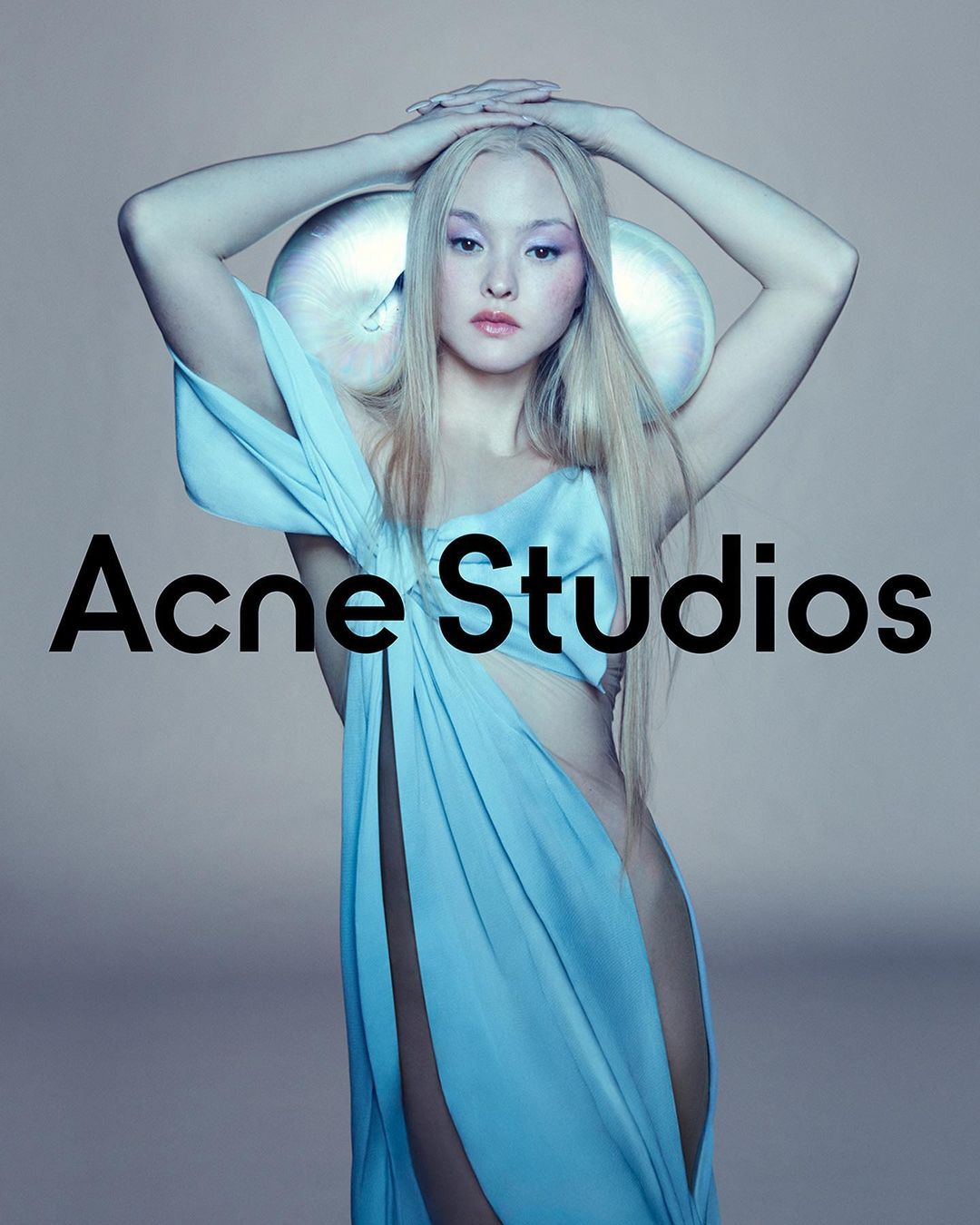 A model poses in a light blue draped garment with futuristic headwear. Acne Studios logo displayed. Studio lighting and soft color palette.