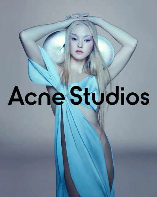A model poses in a light blue draped garment with futuristic headwear. Acne Studios logo displayed. Studio lighting and soft color palette.