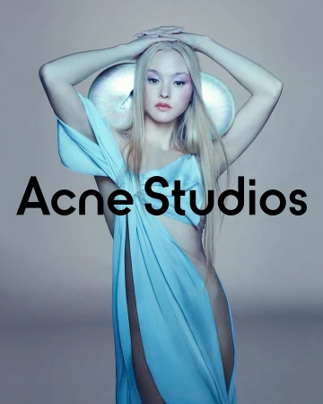 A model poses in a light blue draped garment with futuristic headwear. Acne Studios logo displayed. Studio lighting and soft color palette.