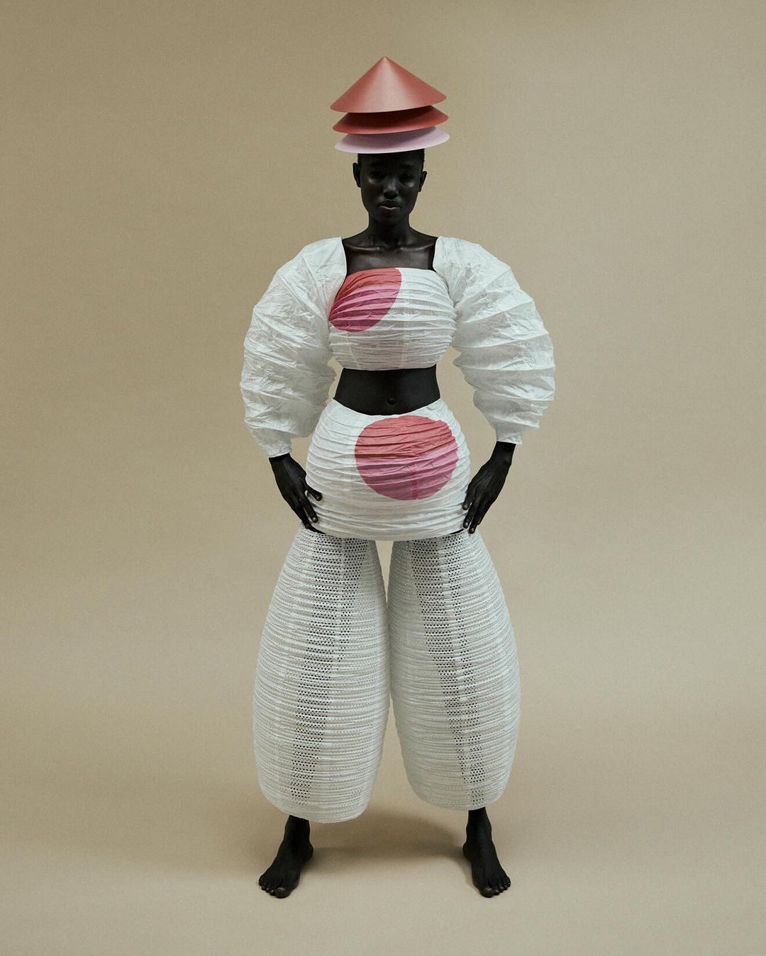 Standing figure in white origami-style fashion with tiered cone hat and pink accents against a neutral background in a studio setting.