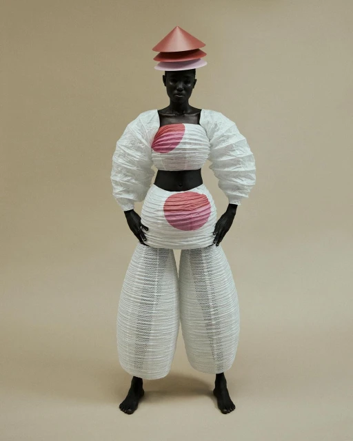 Standing figure in white origami-style fashion with tiered cone hat and pink accents against a neutral background in a studio setting.