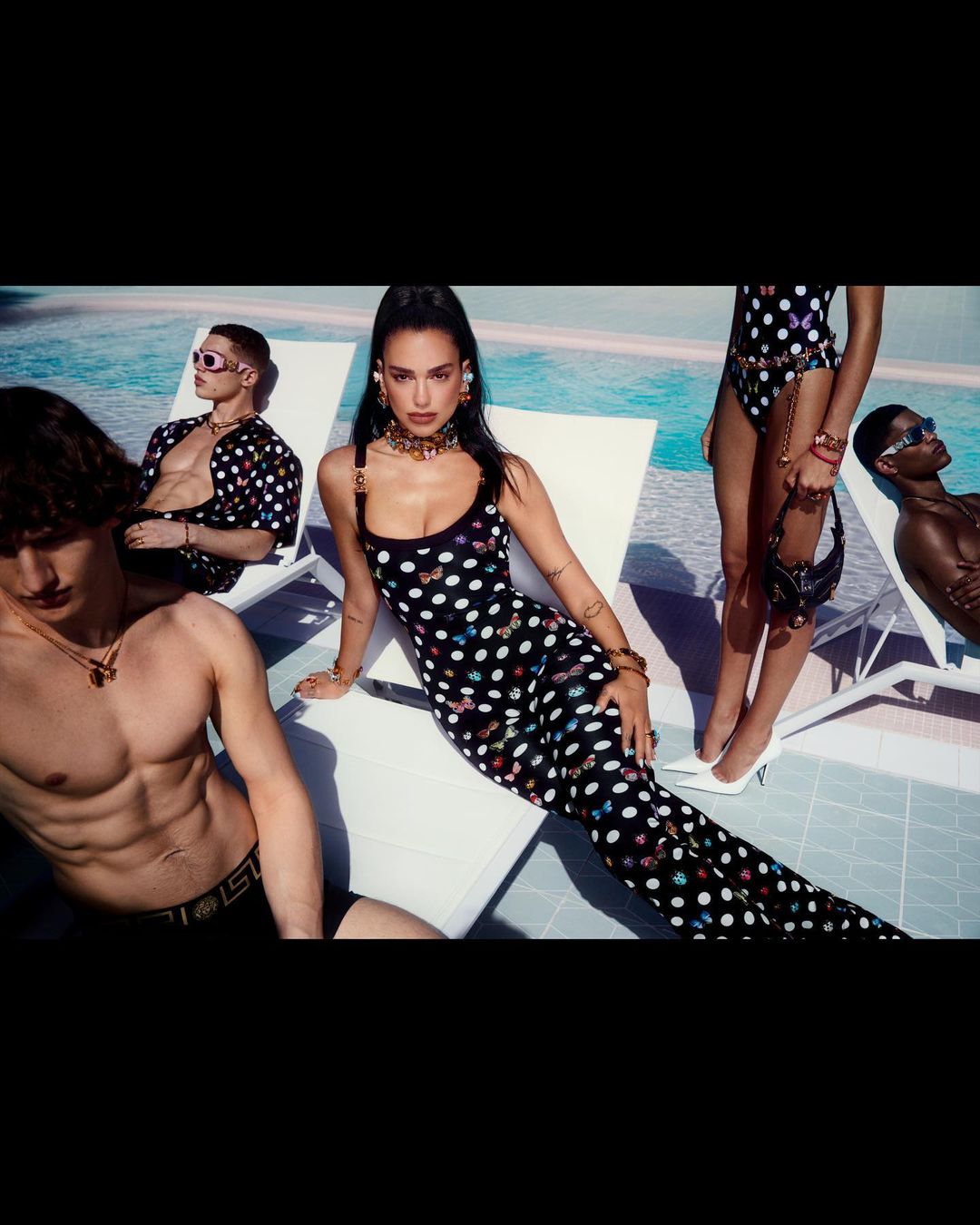 Group of fashion models lounging poolside wearing patterned swimwear and summer attire, accessorized with jewelry and sunglasses.