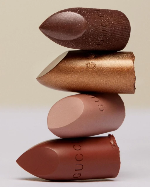 Stacked Gucci lipsticks in bronze, brown, and tan hues create a beauty product still life against a pale backdrop in a vertical studio shot.