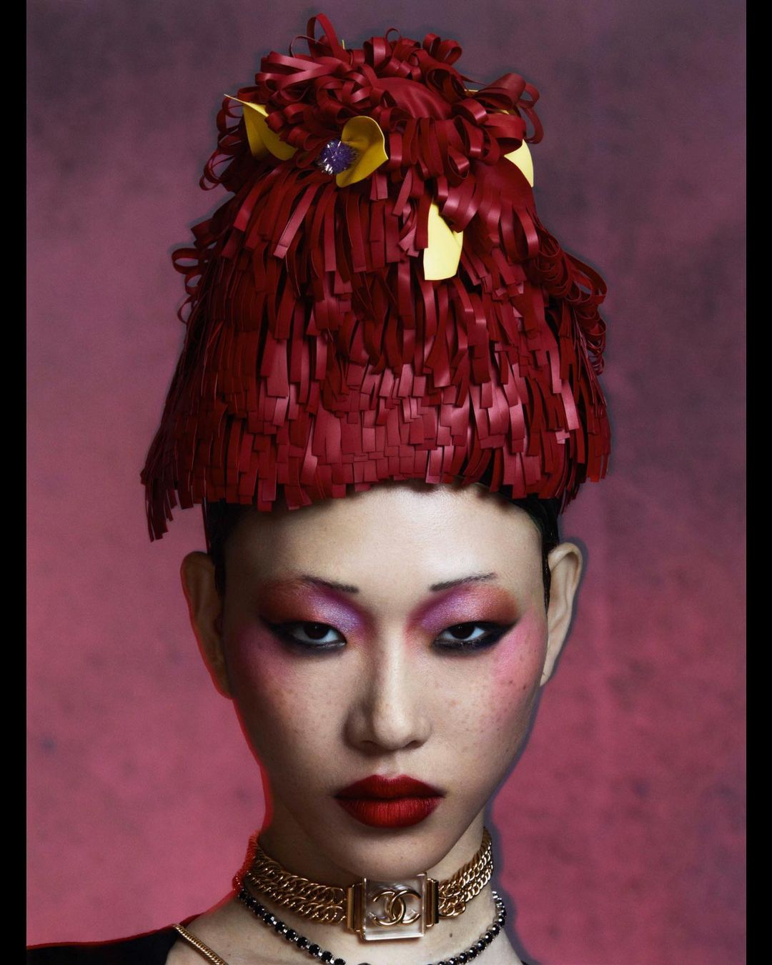 Close-up portrait of a person with pink eye shadow, red lipstick, gold and black necklace, and a red and yellow headdress against a red background.