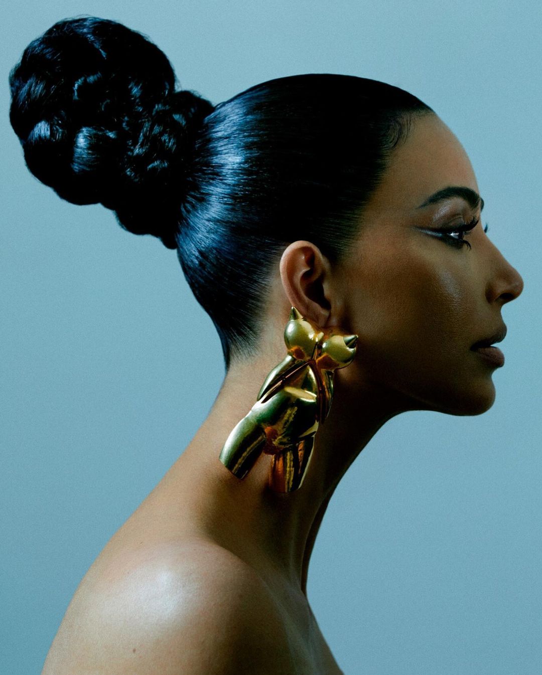 Profile portrait of a woman with her hair styled in a large bun, wearing golden bow earrings, against a pale blue studio background.
