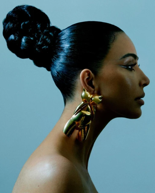 Profile portrait of a woman with her hair styled in a large bun, wearing golden bow earrings, against a pale blue studio background.