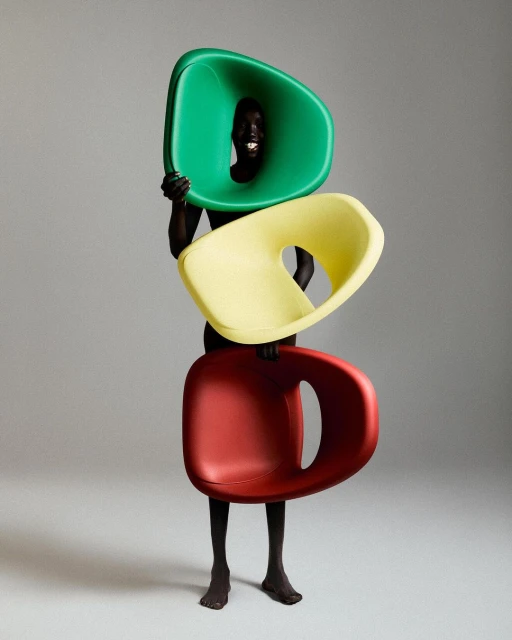 Person holding a tower of red, yellow, and green modern plastic chairs in a photo studio against a plain gray background.