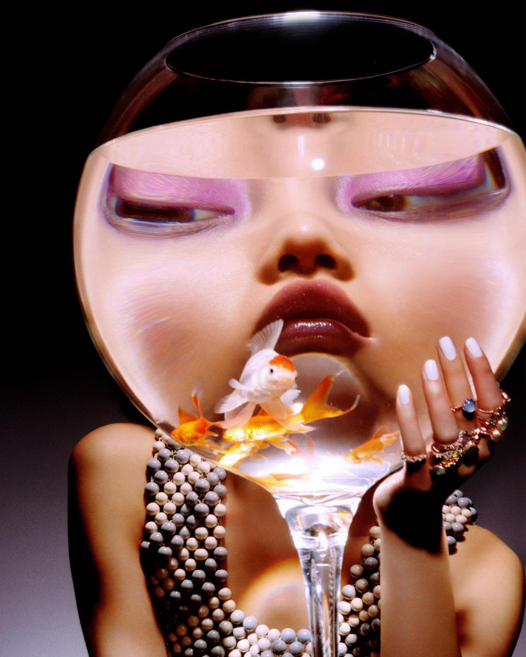 Eye-catching portrait of an adult female wearing a pearl dress, with a fishbowl over her head containing several goldfish. 