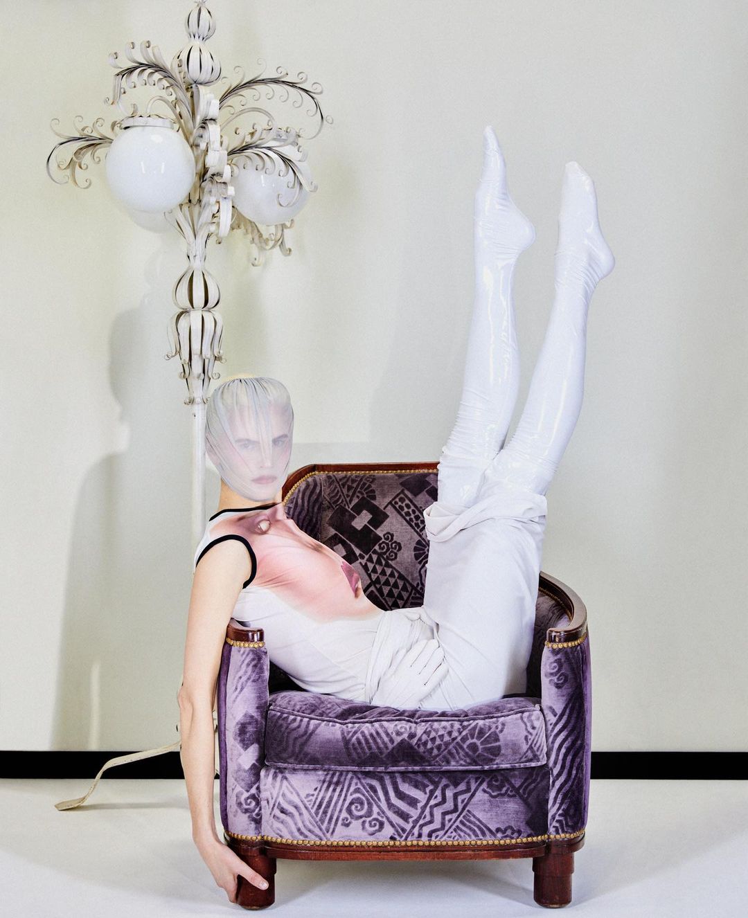 Model in purple patterned chair wearing white leggings and head covering in studio with decorative floor lamp and pale wall.