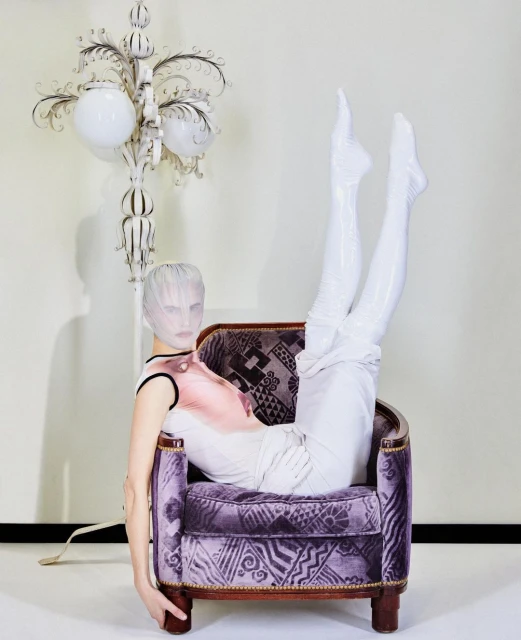 Model in purple patterned chair wearing white leggings and head covering in studio with decorative floor lamp and pale wall.