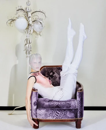 Model in purple patterned chair wearing white leggings and head covering in studio with decorative floor lamp and pale wall.