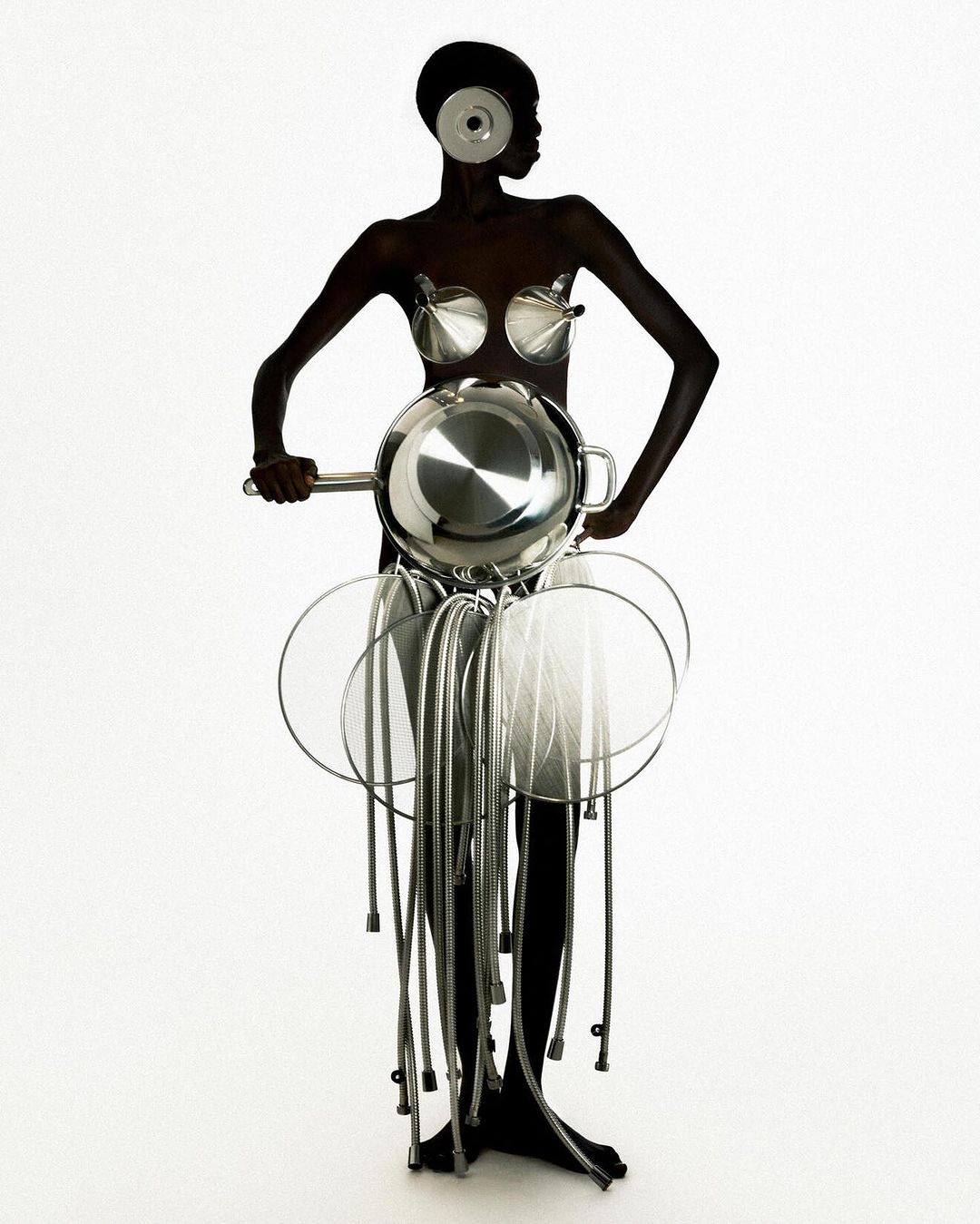 A dark-skinned woman in a conceptual fashion design made of kitchen pots, pans, and utensils poses in a full shot on a white backdrop.