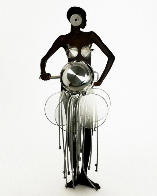 A dark-skinned woman in a conceptual fashion design made of kitchen pots, pans, and utensils poses in a full shot on a white backdrop.