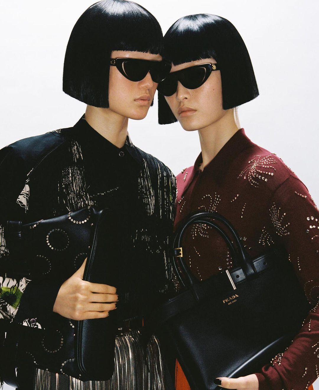 Two women with matching bobs and sunglasses pose closely, each holding a black leather handbag, against a bright, neutral backdrop.