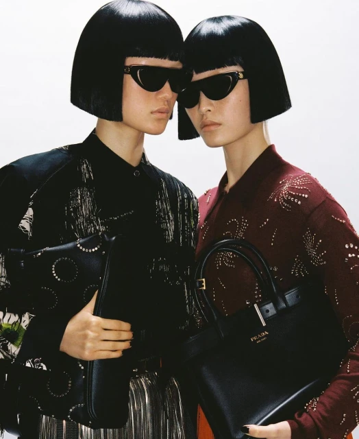 Two women with matching bobs and sunglasses pose closely, each holding a black leather handbag, against a bright, neutral backdrop.