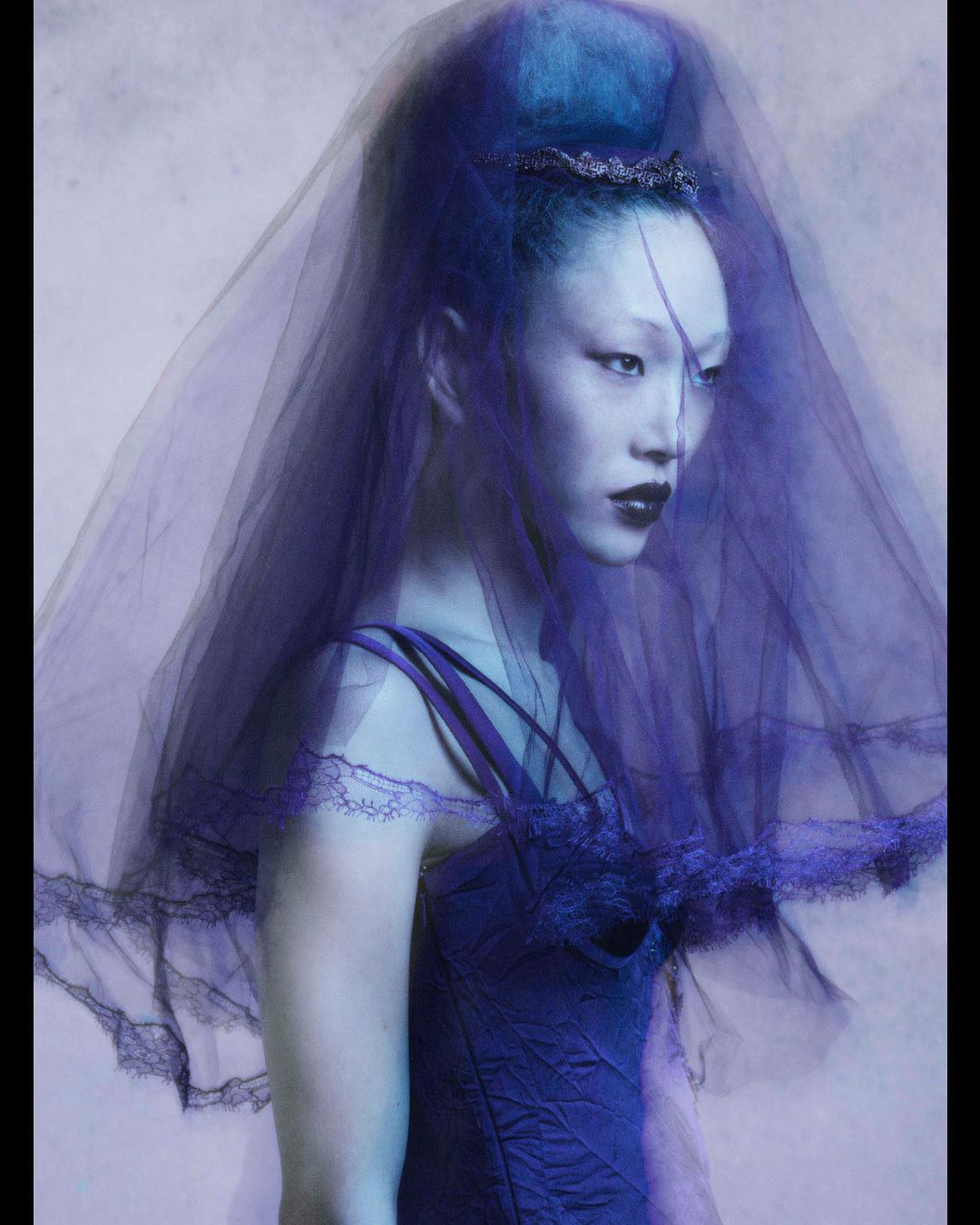 Portrait of a woman with fair skin wearing a blue dress and a veil in front of a light grey backdrop. The lighting is soft and diffused.
