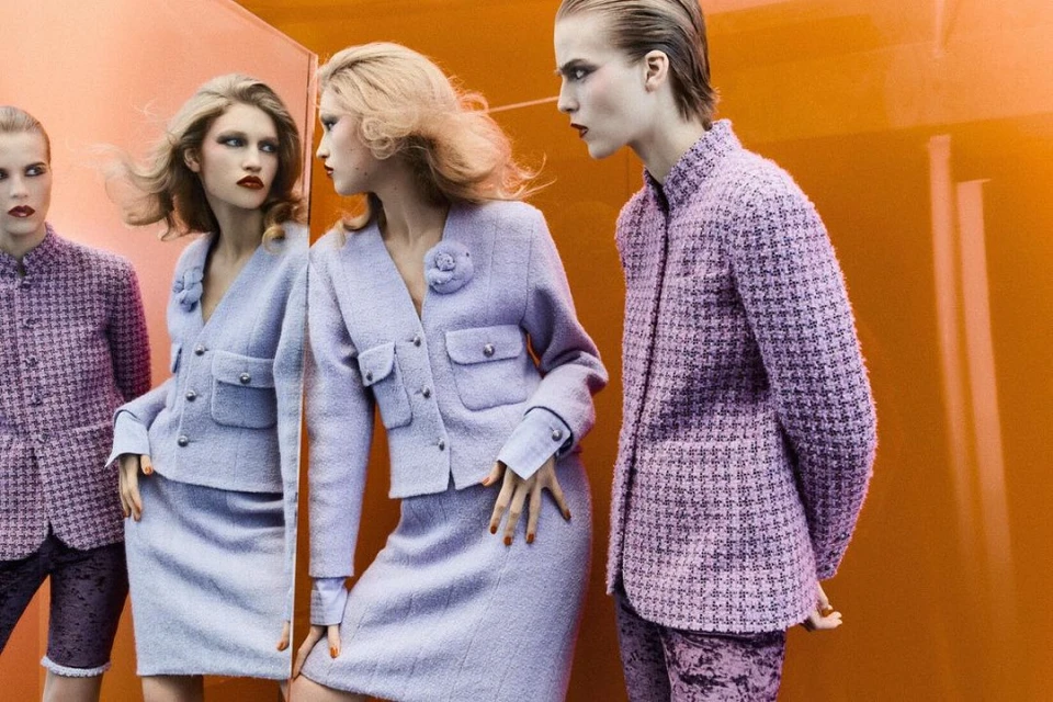 Fashion editorial shot of two women in lilac tweed skirt suits standing against an orange backdrop, one looking in a mirror.