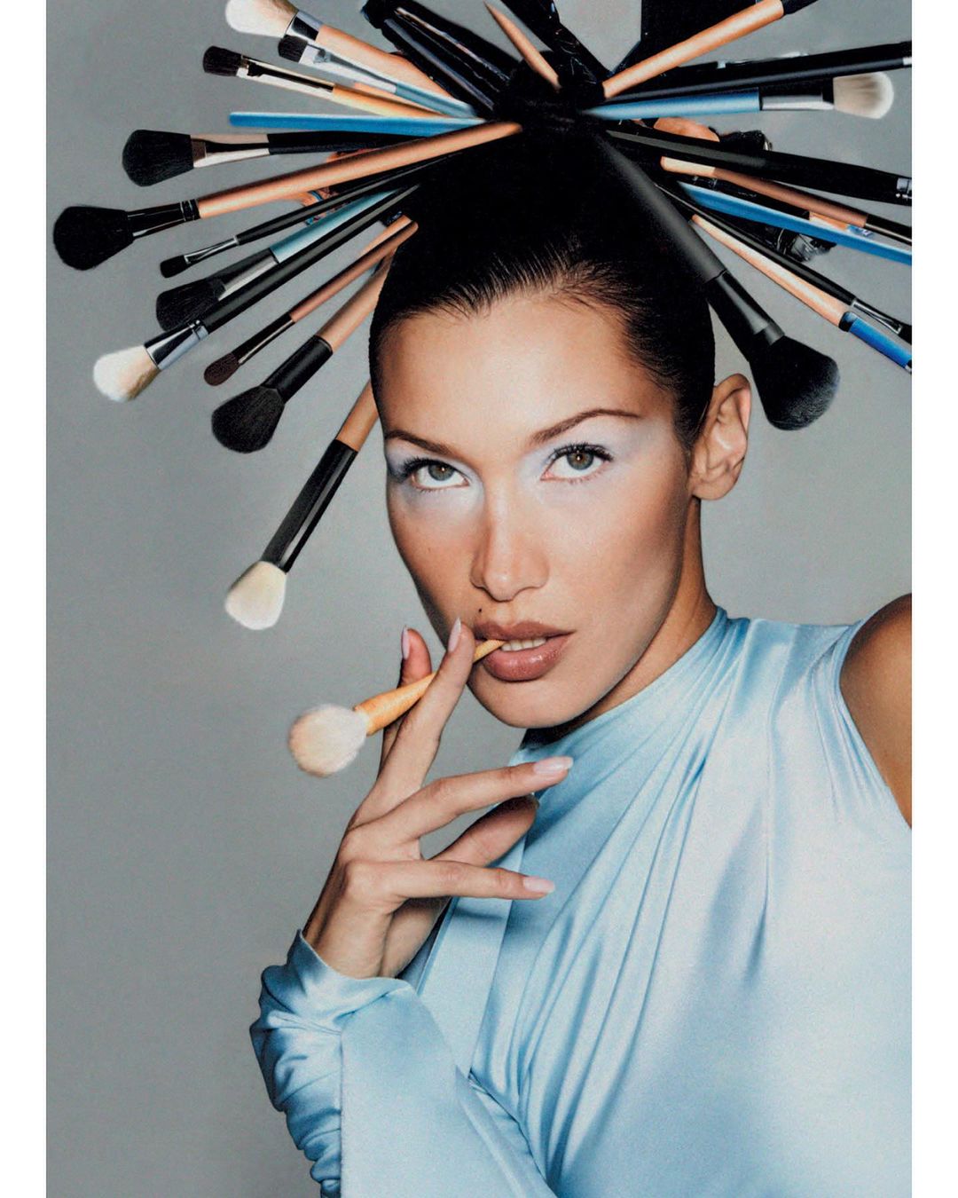 Close-up portrait of a woman with fair skin and light blue eye makeup wearing a light blue gown surrounded by makeup brushes.