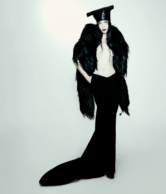 Fashion photo of a woman in a black avant-garde outfit with a dramatic feather stole, posing against a bright, minimalist background.