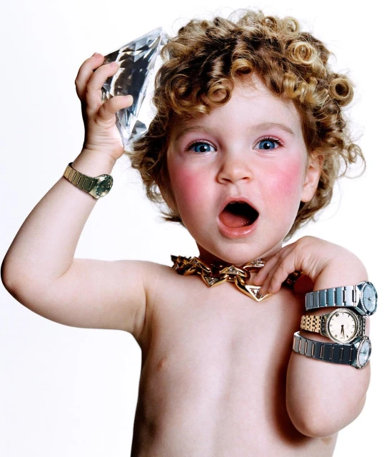 Toddler with curly hair poses with a large diamond prop, wearing multiple wristwatches and a thick gold necklace against a white background.