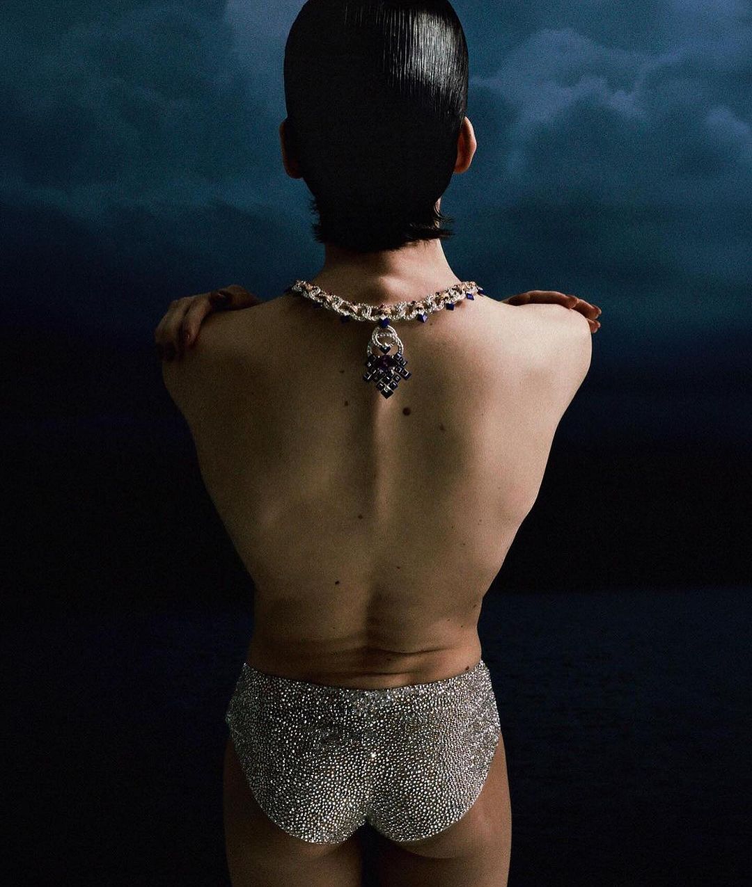 Back view of person with dark hair wearing a necklace and sequined shorts against a dark blue background.