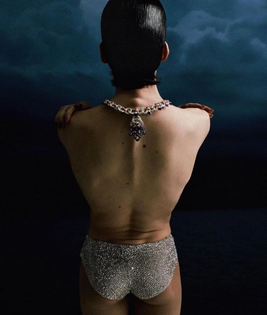 Back view of person with dark hair wearing a necklace and sequined shorts against a dark blue background.