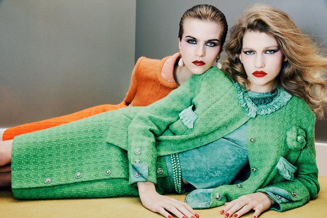 Two women in green and orange suits pose in a fashion editorial, showcasing bold makeup and retro styling against a neutral backdrop.